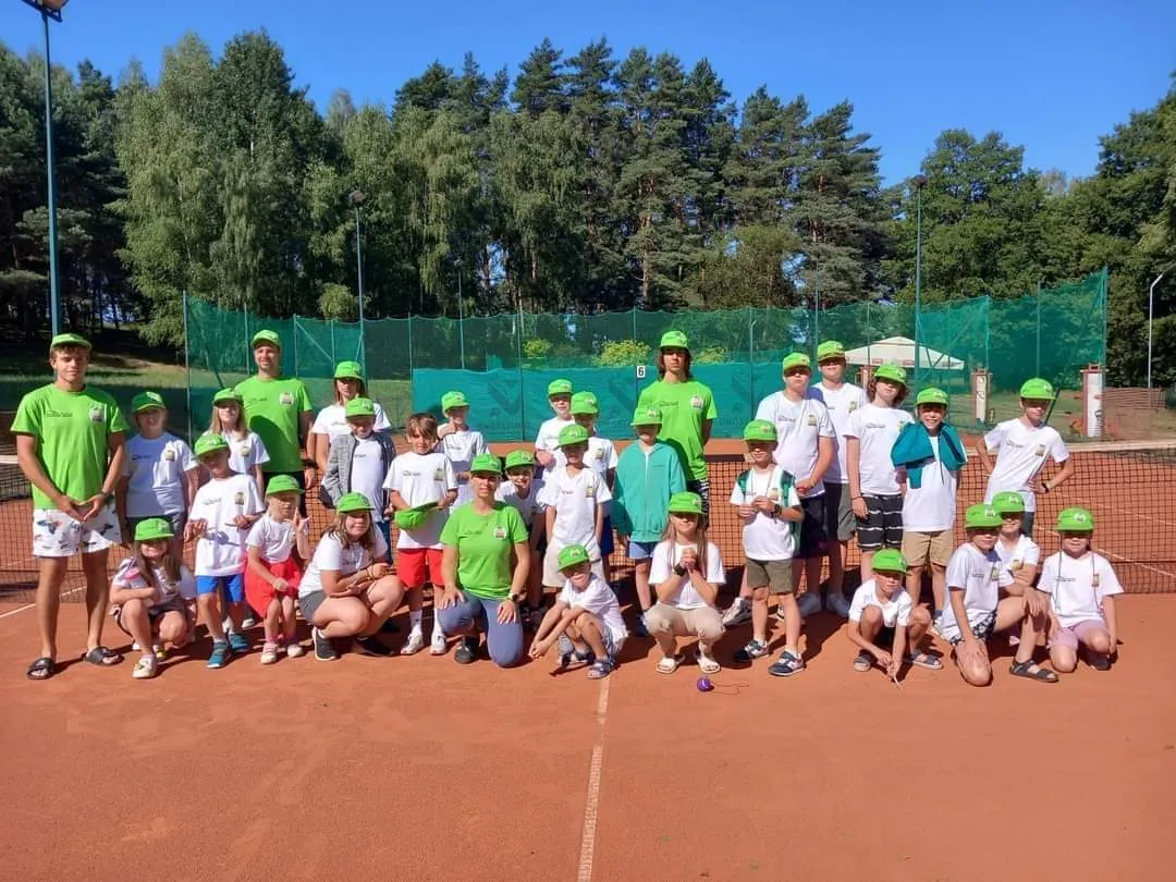 Grupa uczestników obozu z trenerami na korcie ziemnym, w zielonych czapkach Tennis Spot